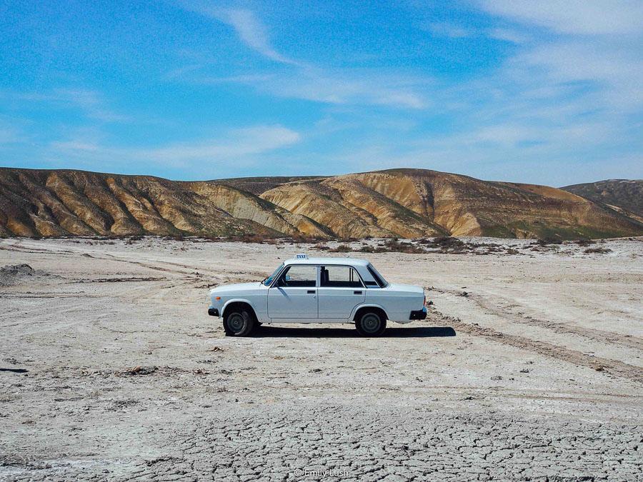 A white car parked on a dry riverbank with brown-coloured mountains in the background.