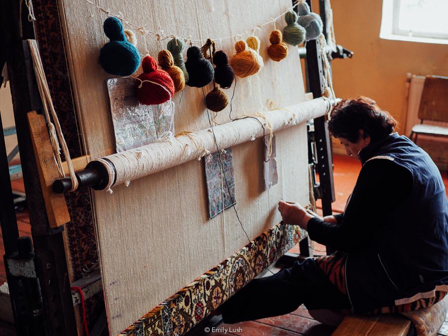 A woman works at a carpet loom with colourful balls of wool.