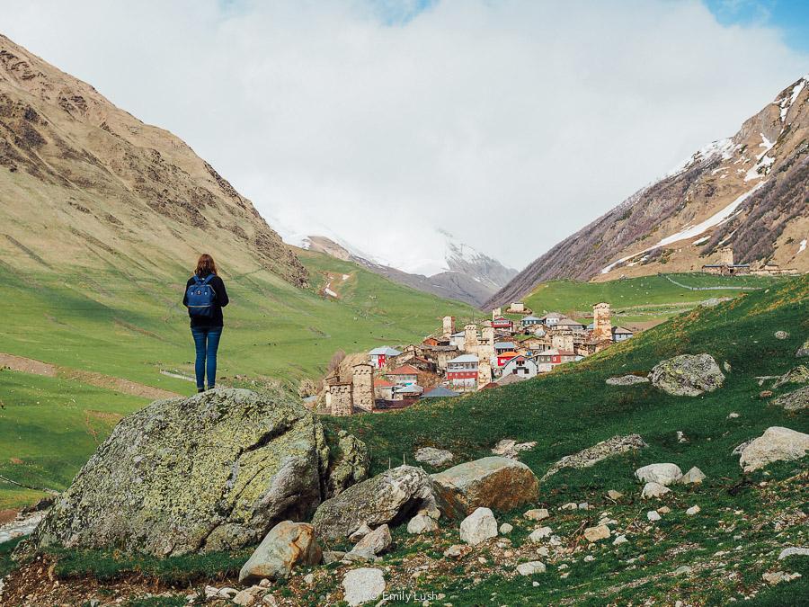 A woman stands on a rock overlooking a small village with stone towers in Svaneti.