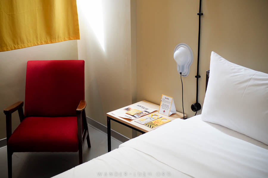 A bed with white linens and a red chair in a hostel room in Tbilisi.
