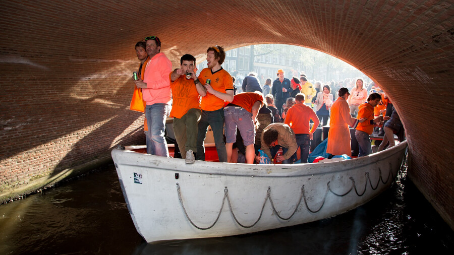 A group of men in orange shirts ride in a boat under a low bridge in Amsterdam.