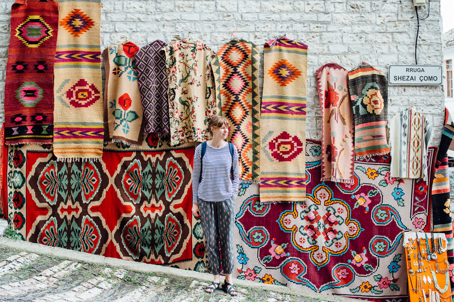 A woman stands in front of a display of brightly coloured carpets in Albania.