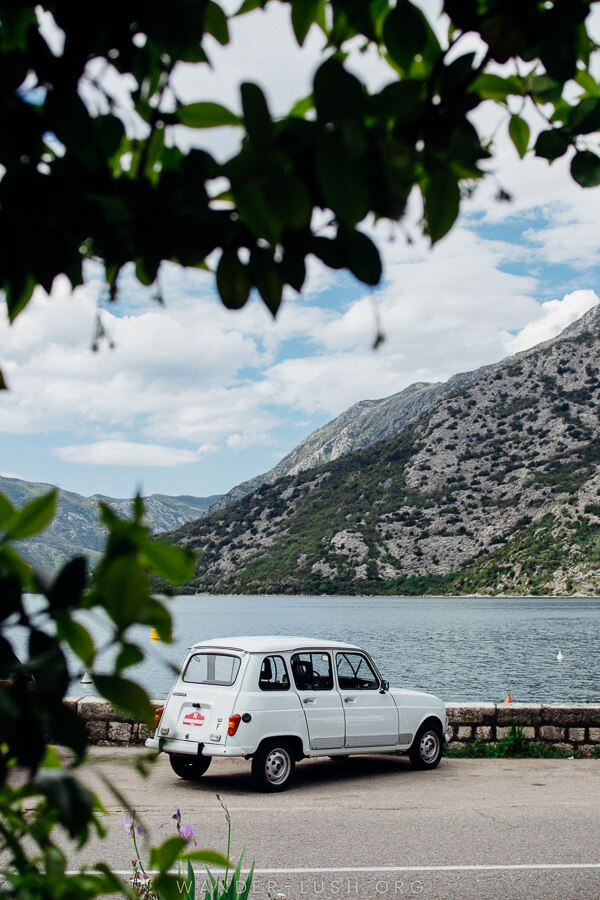 A retro white car parked in front of the ocean with a grey mountain in the background.