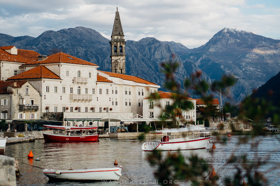 A white Venetian building on a harbour with boats and a stone tower in the background in Perast, Montenegro.