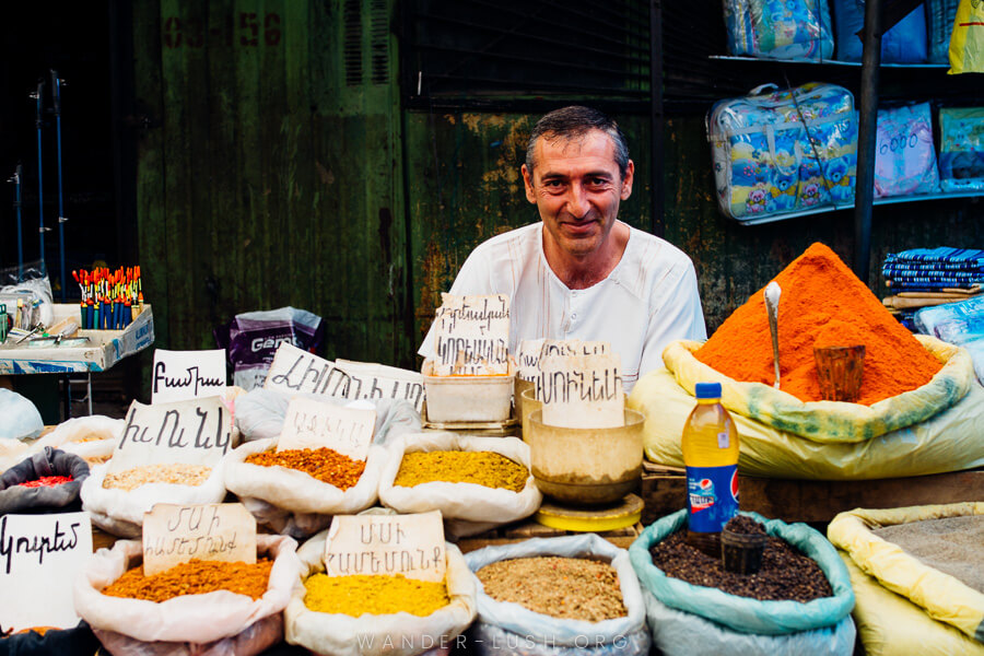 A man in a white shirt sits behind a market table in Gyumri, Armenia selling bags of brightly coloured spices.