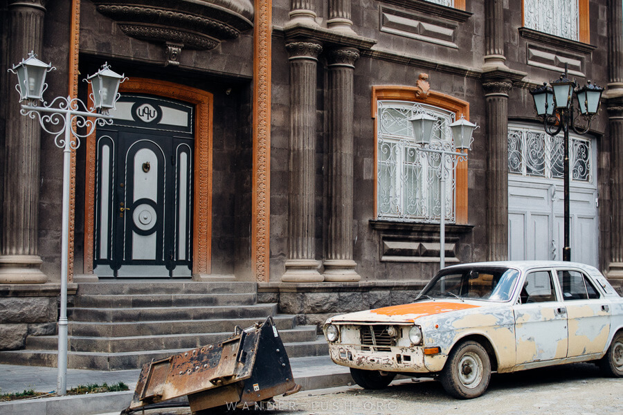 A grey and black stone building with a wrecked white car out the front.