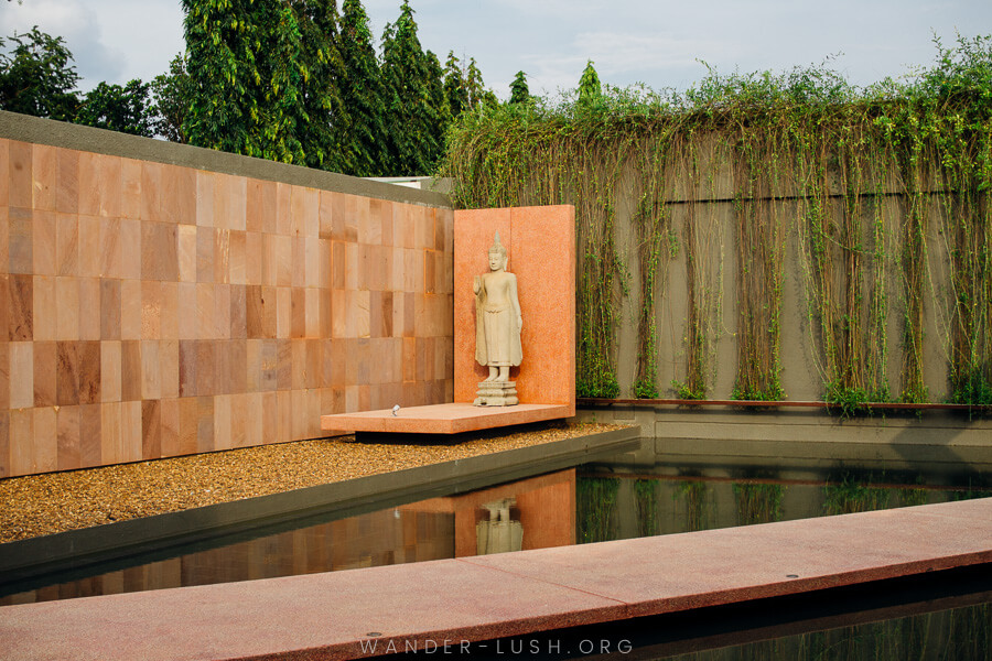 A Buddha statue sits against a pink-rock wall over a shallow pond.