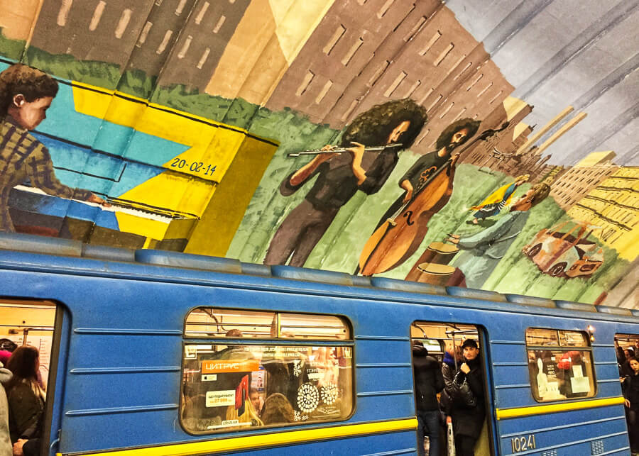 A blue train runs through a metro station in Kyiv, with murals depicting musicians on the roof.