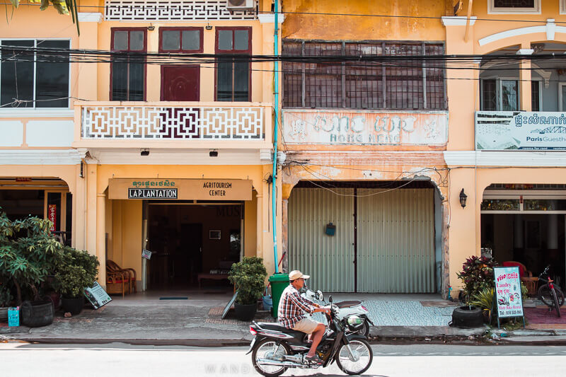 A man rides a motorbike past a row of yellow and white shophouses in Kampot, Cambodia.