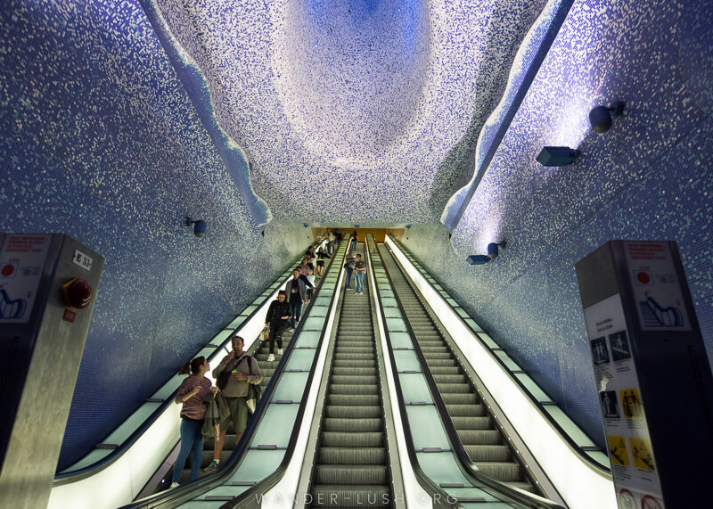 Escalators inside an underground metro station at Toledo in Naples, with blue and white tiles on the walls and ceiling.
