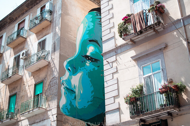 A blue face peeks out from between apartment buildings - street art in Naples.