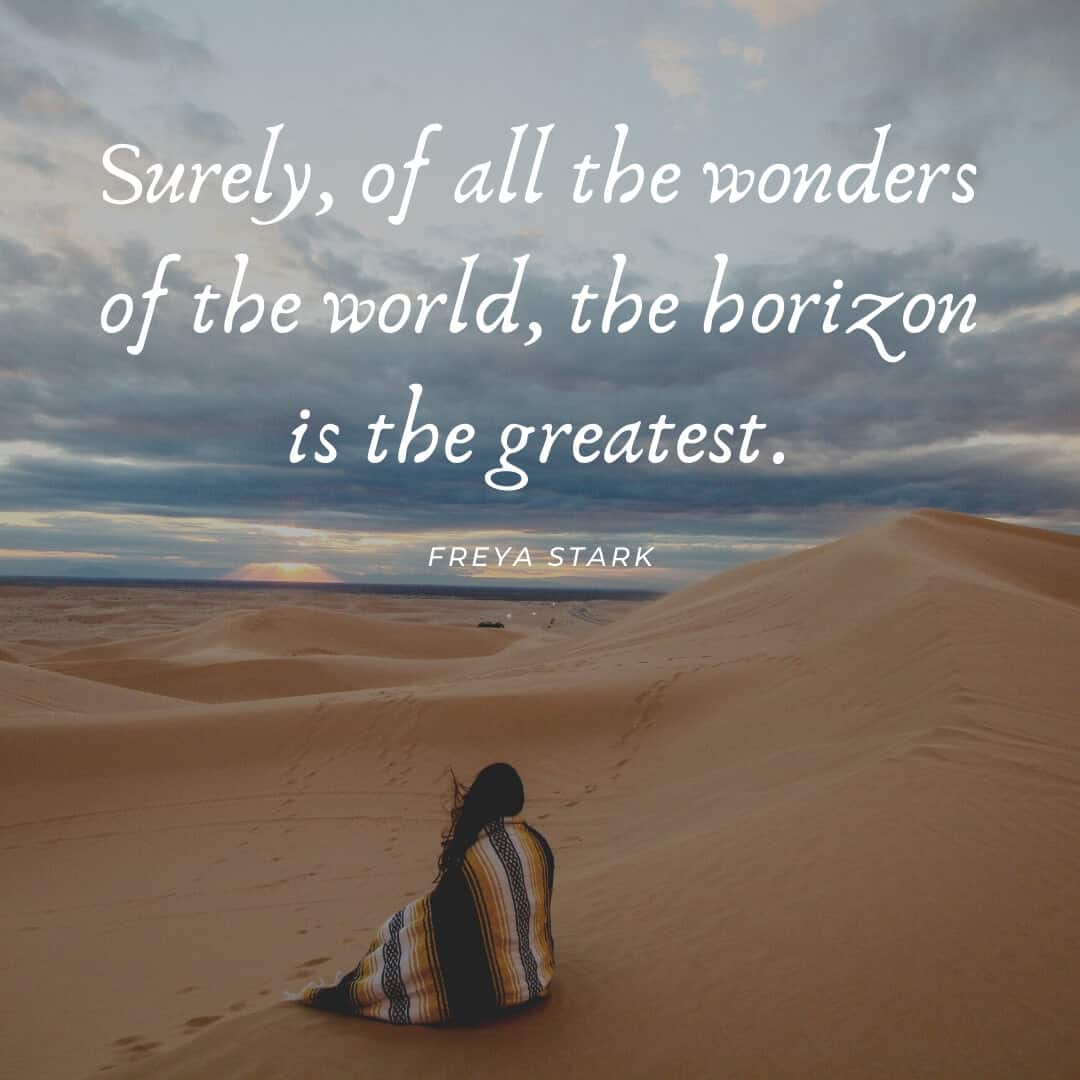 A woman wrapped in a blanket sits on a sand dune and looks out to the horizon.