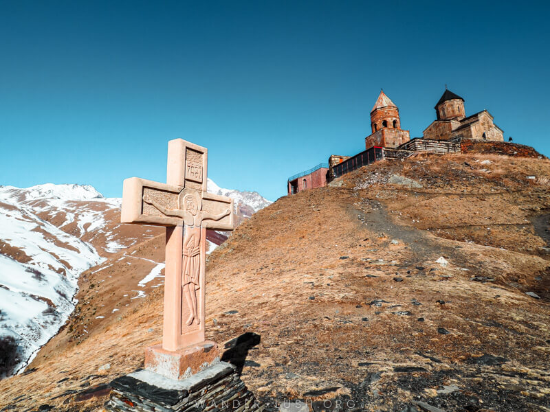 A stone church and a cross atop a snowy mountain in Georgia.