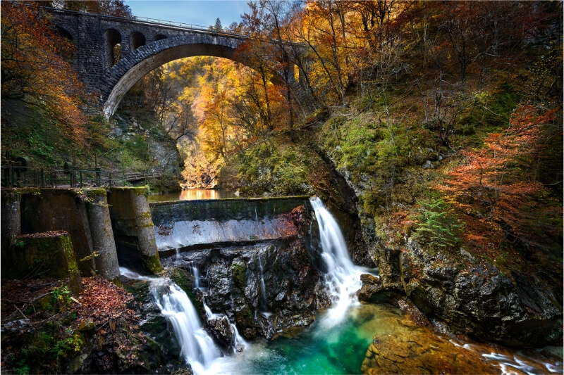 A beautiful stone bridge over a dam surrounded by autumn foliage.