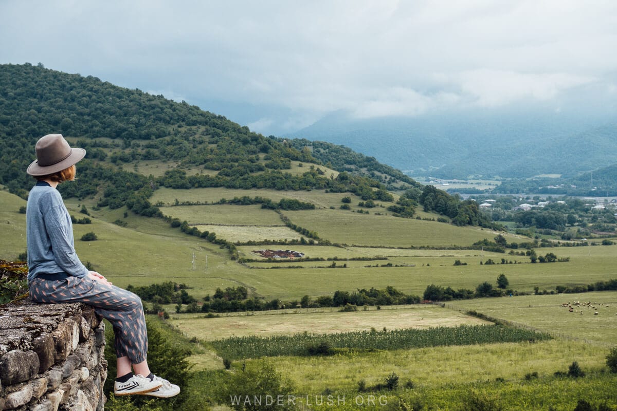 A woman sitting on a fortress wall overlooking green pastures and mountains in the countryside of Georgia.
