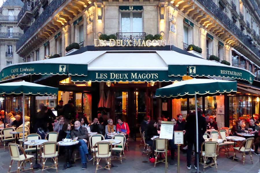 People drink coffee on sidewalk tables at Les Deux Magots in Paris.