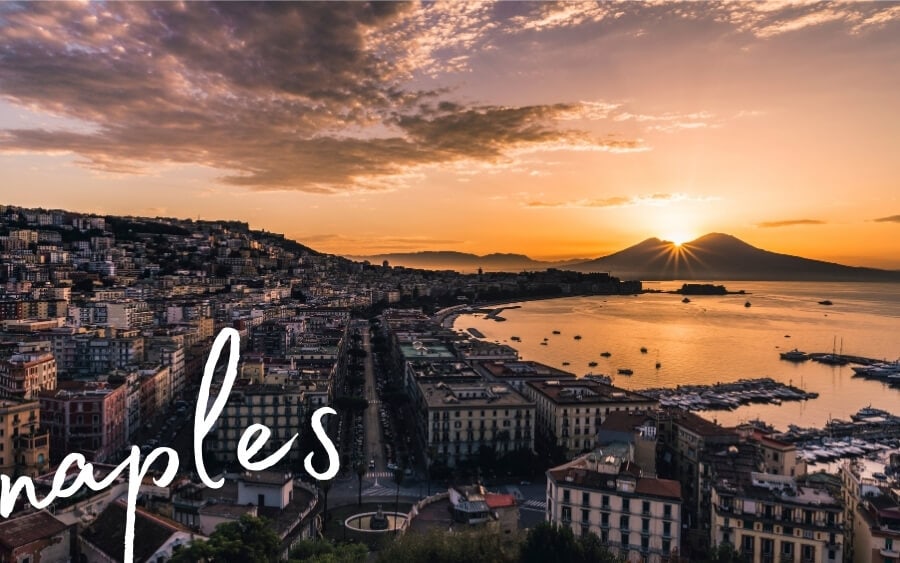 The city of Naples at dusk, with the sun setting over a volcanic mountain in the background.