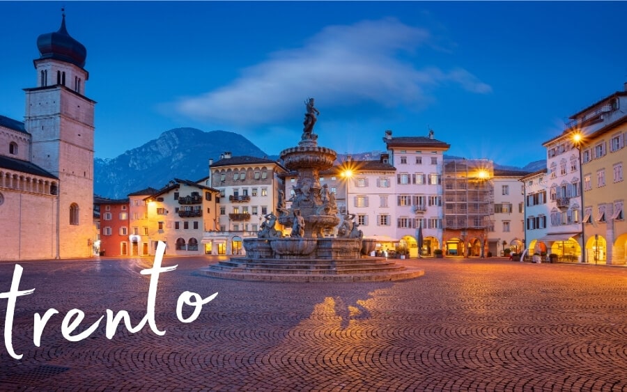 Trento, Italy by night, with lights illuminating buildings around a main square and fountain.