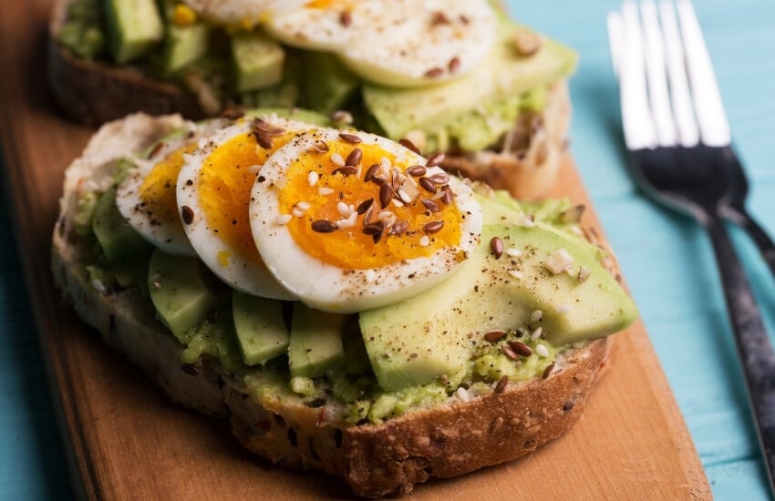 A piece of grain toast topped with sliced avocado and boiled eggs.