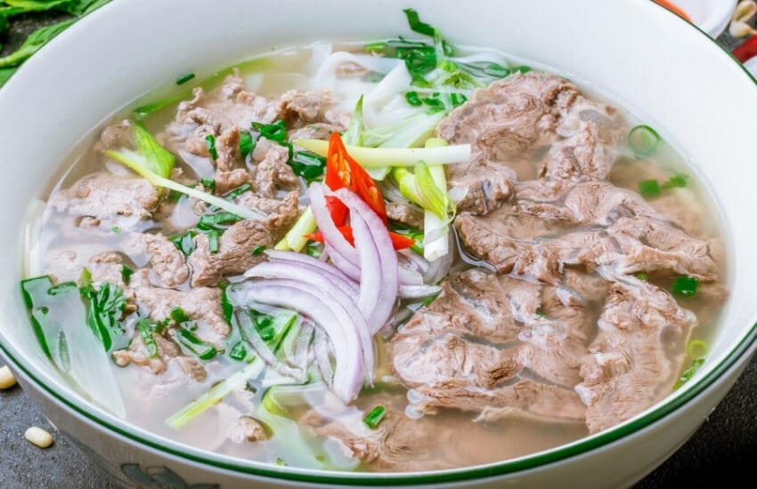 A bowl of pho soup, with sliced beef and onions.