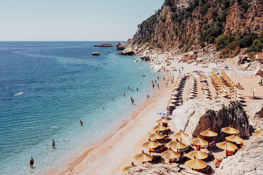 People sit under umbrellas on a white sandy beach in Montenegro.