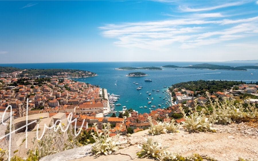 Peering over a stone wall to view Hvar, an old town and bay with islands visible on the horizon.