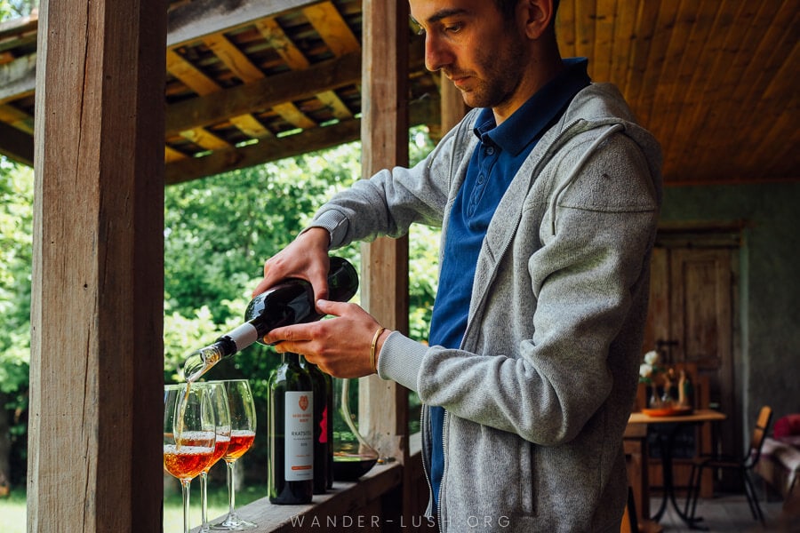 A young man dressed in a grey jacket pours amber wine into three glasses.