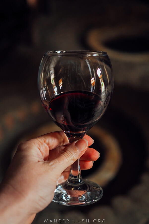 A hand holds a glass of red wine against a clay qvevri.