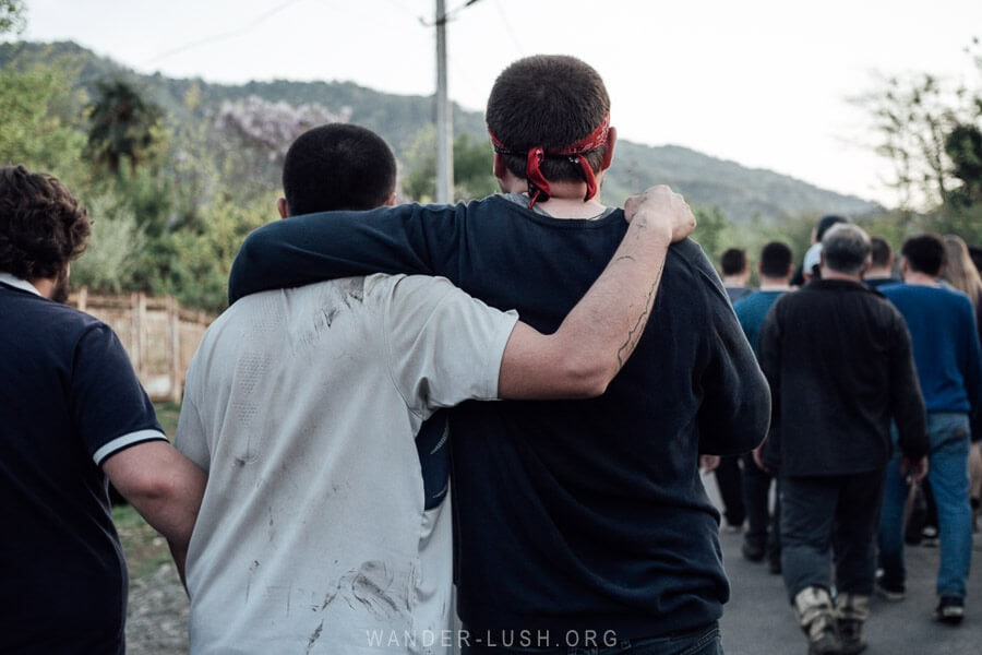 Two young Georgian men with their arms around each other's shoulders walking towards a village at the end of the Lelo Burti Easter festival in Guria.