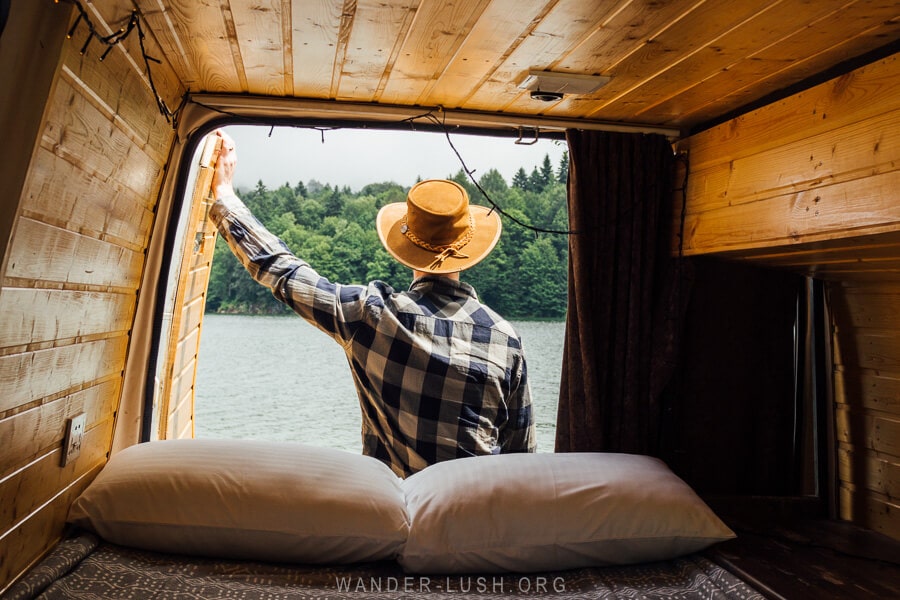 A man in a hat stands at the back of a campervan overlooking Shaori Reservoir in Racha, Georgia.