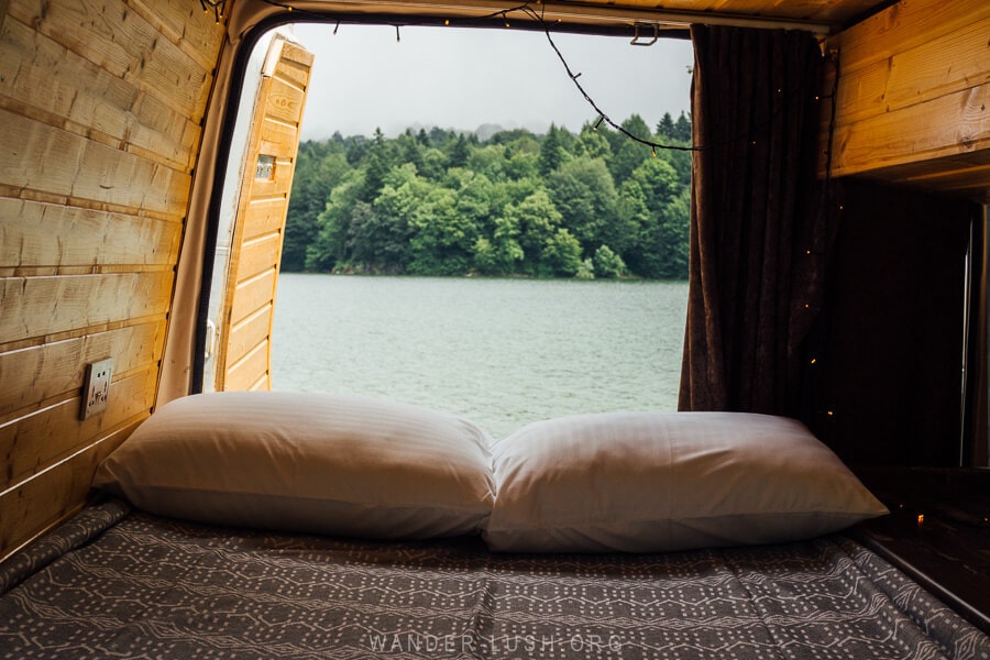 Inside a campervan, a queen sized bed looking out onto a lake and forest.