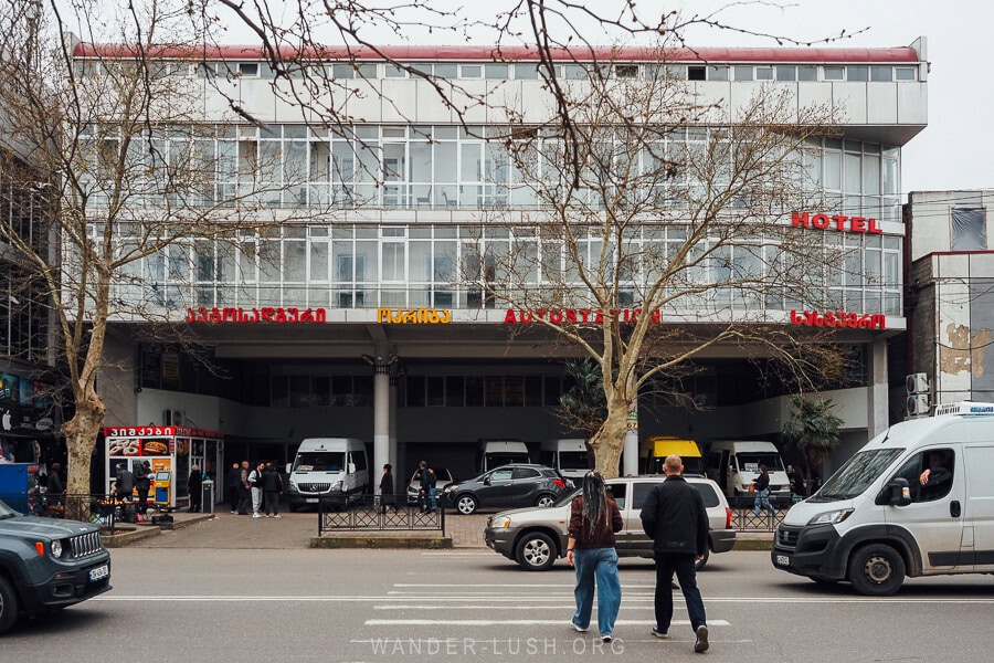 Two people cross the road between cars and vans to reach a bus station on the main highway in Kutaisi, Georgia.