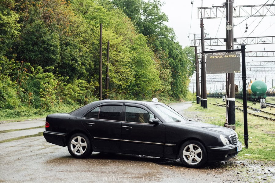 A black car with a taxi sign on its roof parked in front of an electronic bus timetable at Rioni Railway Station near Kutaisi, Georgia.