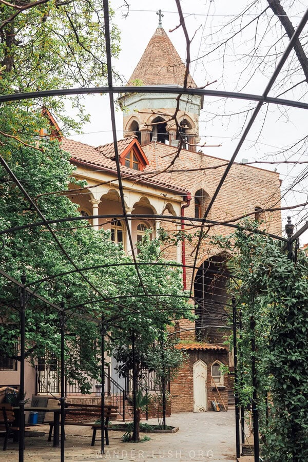 View of a brick gate topped with a crucifix from between the bars of a metal arbour, with spring vines creeping everywhere.