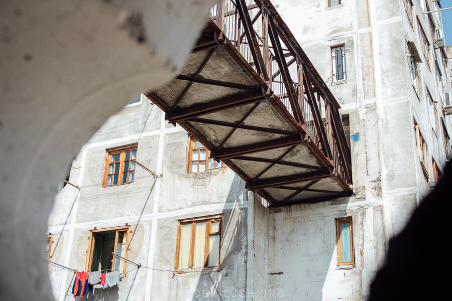 A rusty metal bridge running between two concrete apartment blocks in Tbilisi.