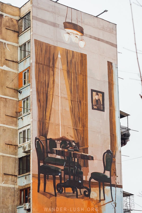A street art mural covers the entire side of a tall apartment building in Varketili district, Tbilisi, depicting the interior of Ilia Chavchavadze's house.