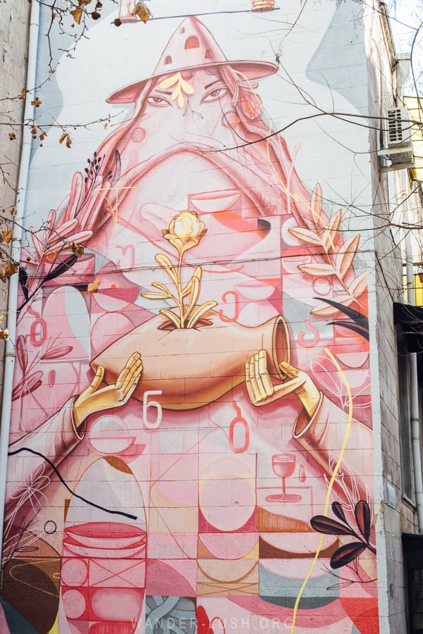 A street art mural in Tbilisi, Georgia depicts a pink figure holding a clay qvevri vessel used for winemaking.