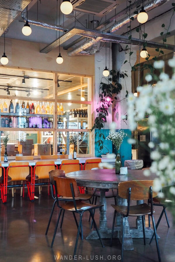 An industrial space with painted walls and exposed concrete retrofitted as a trendy restaurant, with wine stacked on wall shelves and leafy plants.