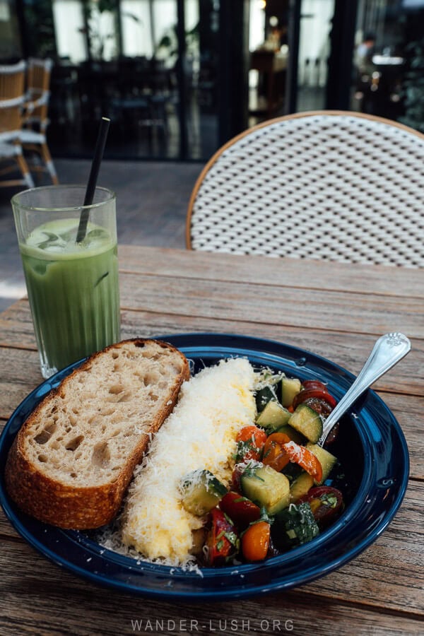 A plate of fresh salad and scrambled eggs served with a slice of bread and a vibrant Matcha latte at a breakfast cafe in Tbilisi, Georgia.