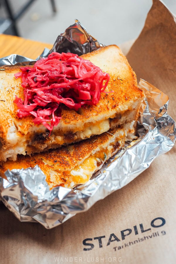 A gourmet cheese toasty topped with pickles spread out on a brown paper bag atop a sidewalk table at a breakfast spot in Tbilisi, Georgia.