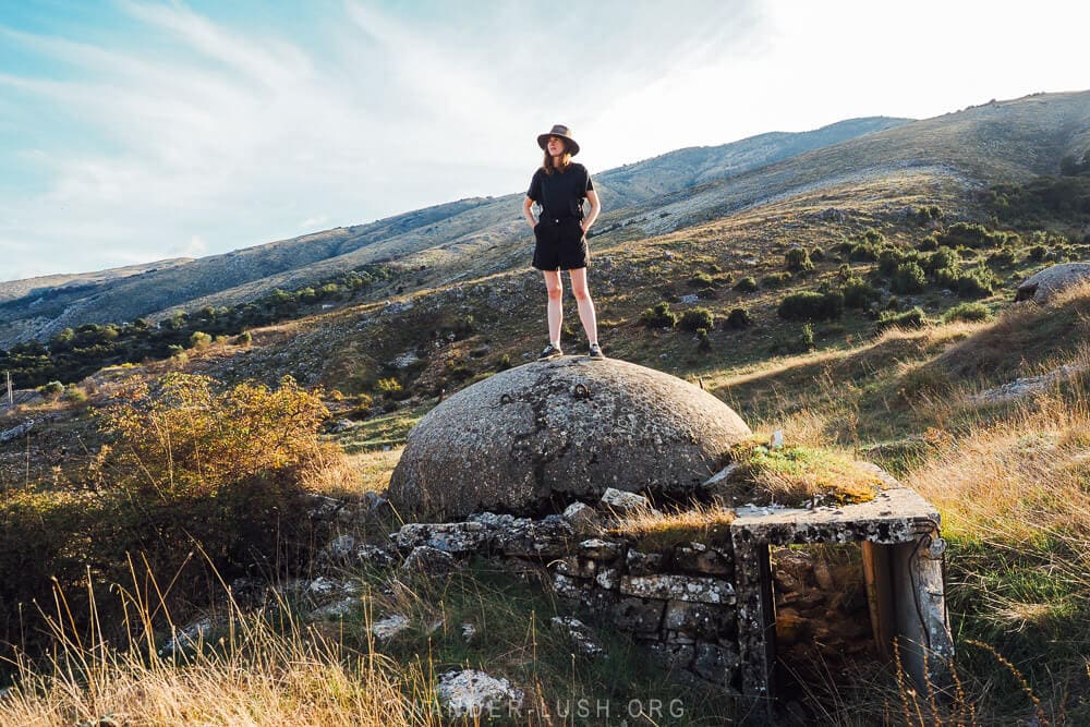 A woman standing on top of a concrete bunker in the middle of a field in Albania at sunset.