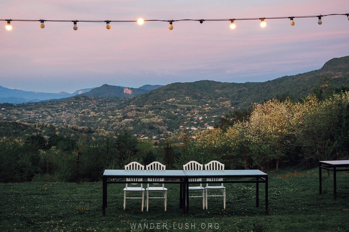 A table with four white chairs set beneath a row of fairy lights on a grassy hill in Kutaisi, with a view of the city below at sunset.