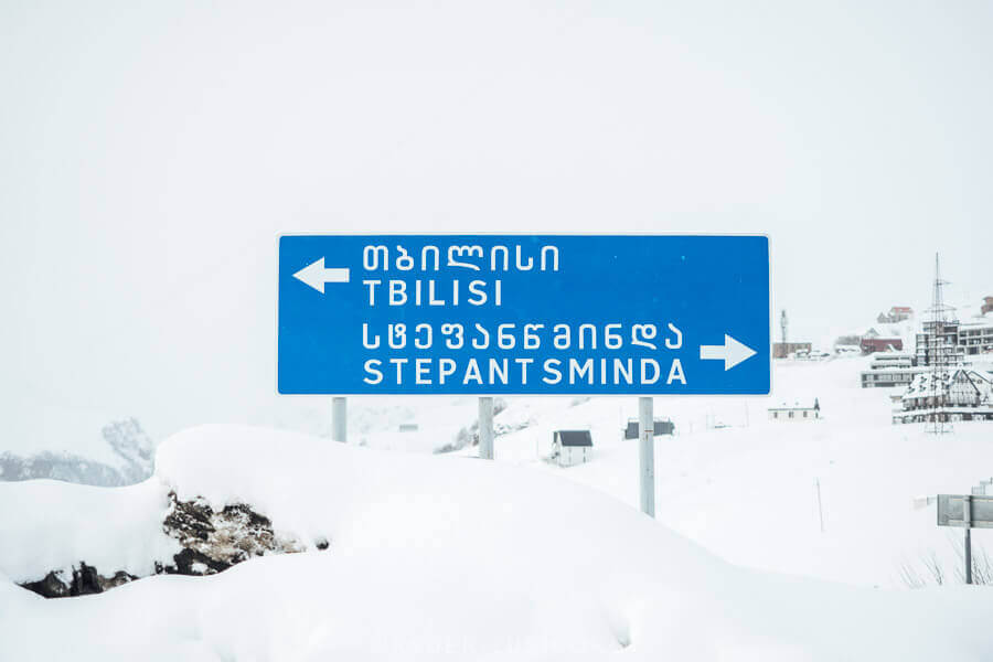 A blue road sign reading Tbilisi and Stepantsminda in English and Georgian in the ski resort of Gudauri.