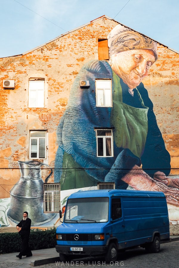A street art mural in Kutaisi, Georgia with a blue van parked in front and a priest walking by.