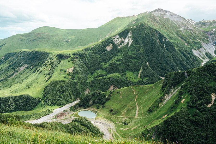 A verdant valley in the Greater Caucasus mountains, with a tiny round blue reservoir at the bottom.