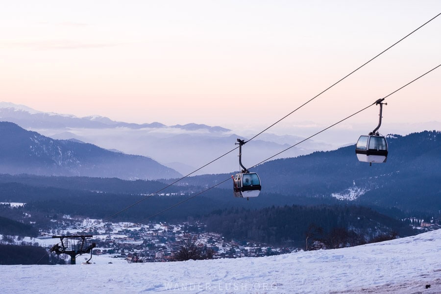 Two modern gondolas sail above the ski resort of Bakuriani in Georgia in winter, with the mountains and sky painted shades of purple during sunset.