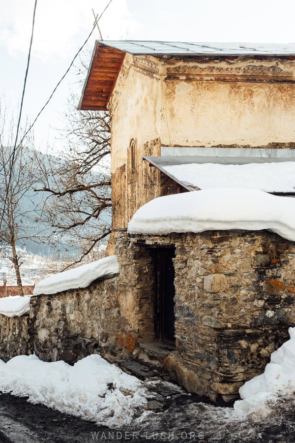 An ancient stone church in Laghami, Mestia, Georgia, covered in snow, featuring a weathered wooden door and traditional architectural details.