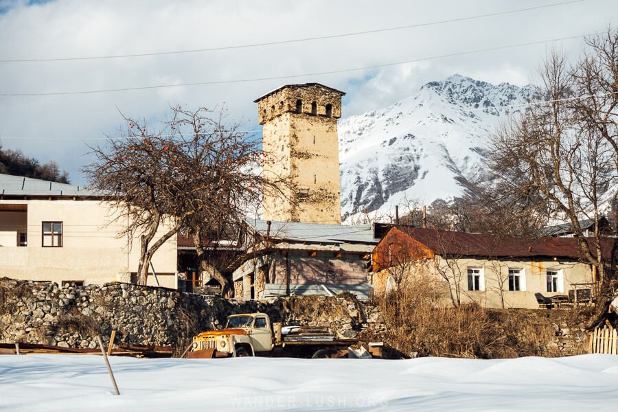 A traditional Svan tower in Laghami, Mestia, Georgia, surrounded by rustic houses and an old yellow truck, with snow-covered mountains in the background.