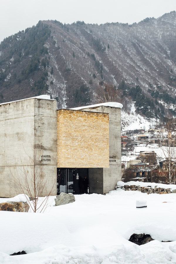 The modern concrete and brick exterior of the Svaneti Museum in Mestia, Georgia, surrounded by snow, with mountains and the town visible in the background.