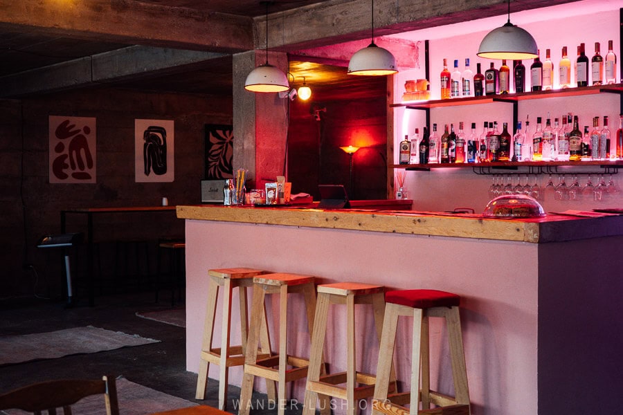 Cozy bar at Twin Peaks in Mestia, Georgia, featuring a rustic-modern design with wooden stools, a pink-lit liquor shelf, and abstract wall art.